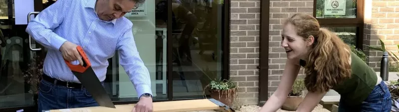 Two volunteers, an older man and a younger woman, saw a plank in the community garden